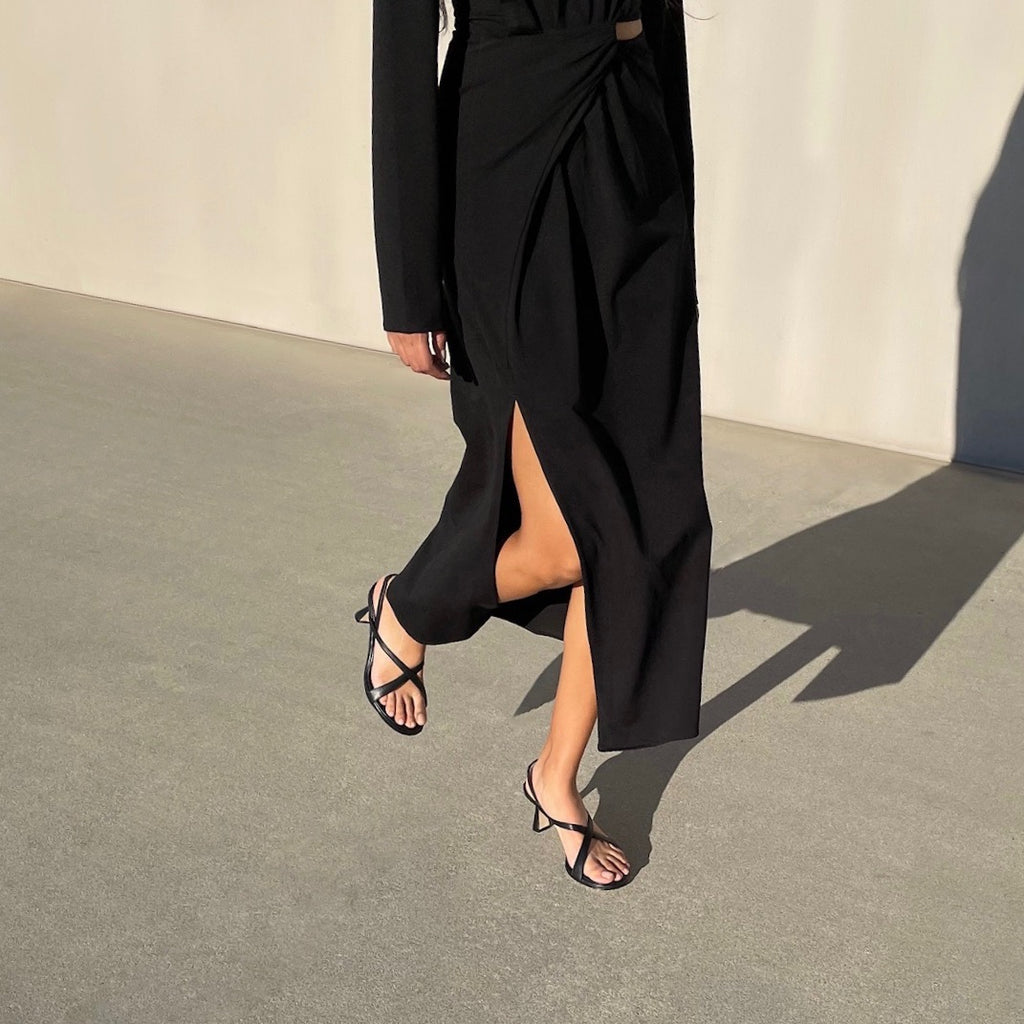 Neil J Rodgers Yin sandal in black nappa leather with a round toe, minimal criss cross instep straps, 60mm heel, and slingback strap is worn with a black long sleeve midi dress.