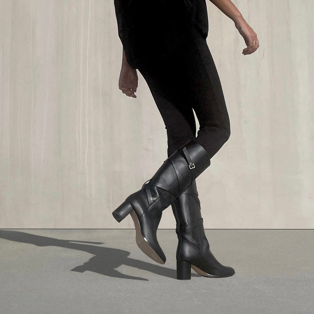BLACK LEATHER MID HEEL KNEE BOOTS WITH STRAP DETAIL ON MODEL WEARING BLACK LEGGINGS IN MINIMAL URBAN CONCRETE SUNNY EXTERIOR