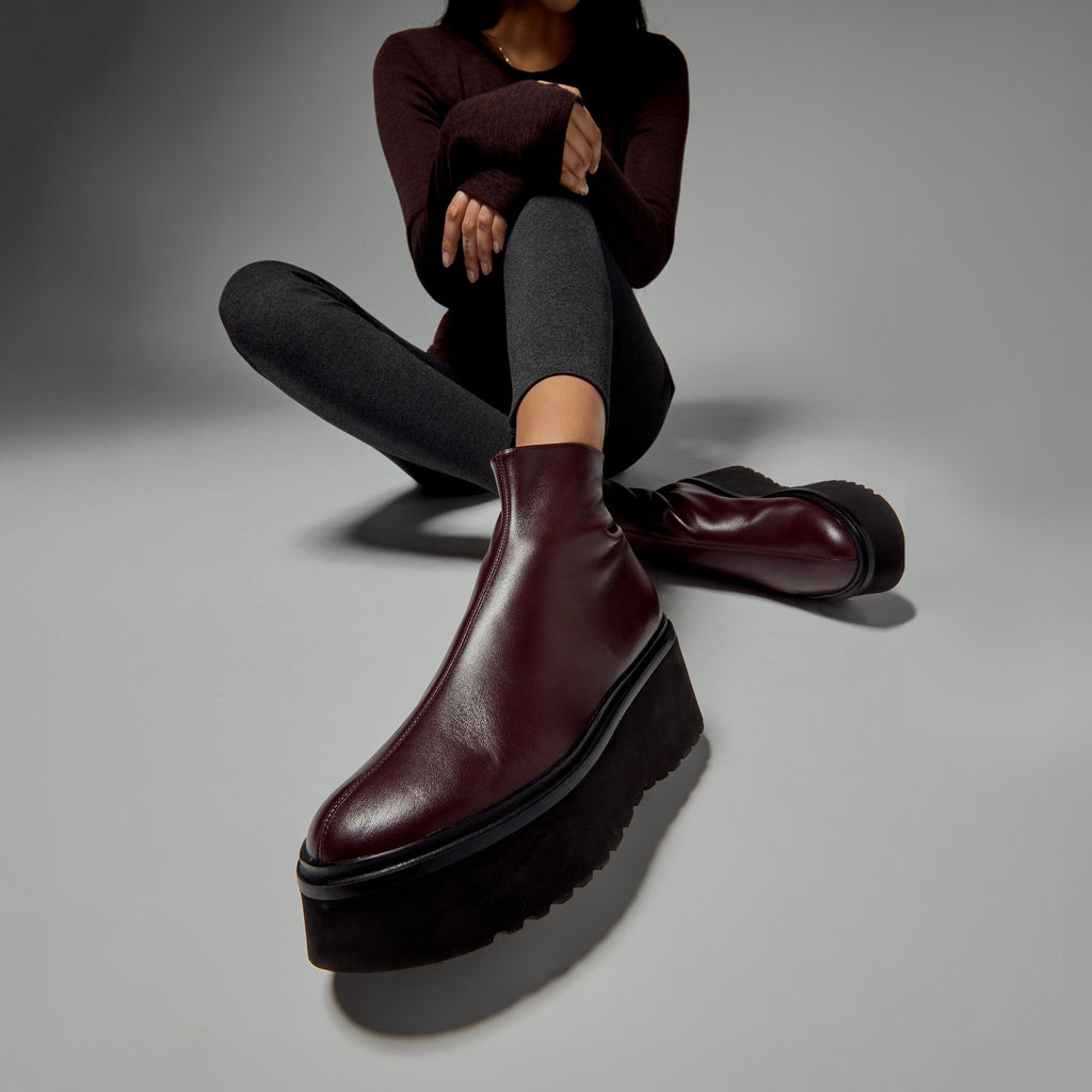 Model wearing platform ankle boots in burgundy leather with black leggings and a thick grey sweater in a minimal urban environment.