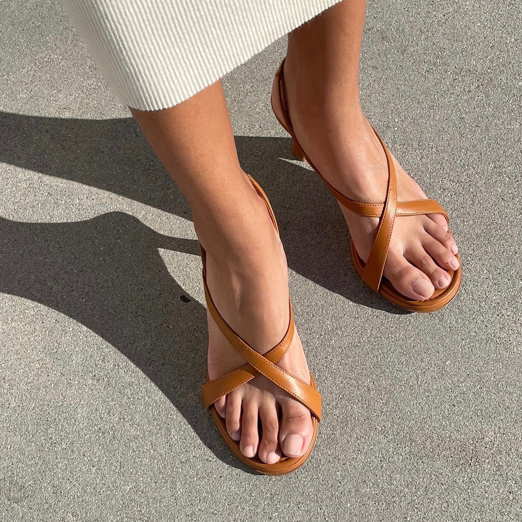 Neil J Rodgers Yin sandal in cognac nappa leather with a round toe, minimal criss cross instep straps, 60mm heel, and slingback strap is worn with a ribbed midi length sweater dress in ivory.