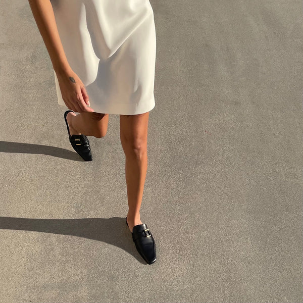 Neil J Rodgers black Andi backless loafer with an angled square toe, minimal stitching, and gold hardware detail in soft Italian nappa leather worn with a short white dress.