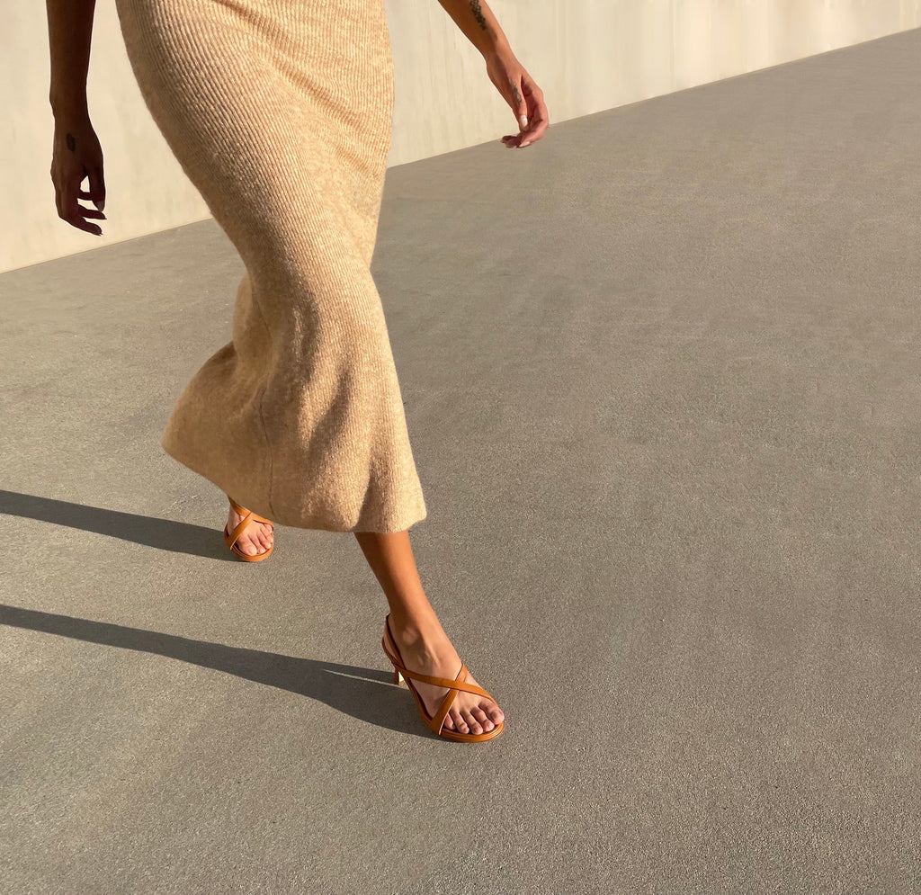 Neil J Rodgers Yin sandal in cognac nappa leather with a round toe, minimal criss cross instep straps, 60mm heel, and slingback strap is worn with a beige midi length dress.