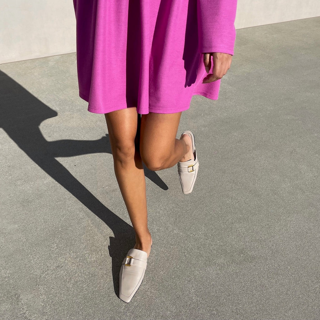 Neil J Rodgers off-white Andi backless loafer with an angled square toe, minimal stitching, and gold hardware detail in soft Italian nappa leather worn with a short, long sleeve fuchsia dress.