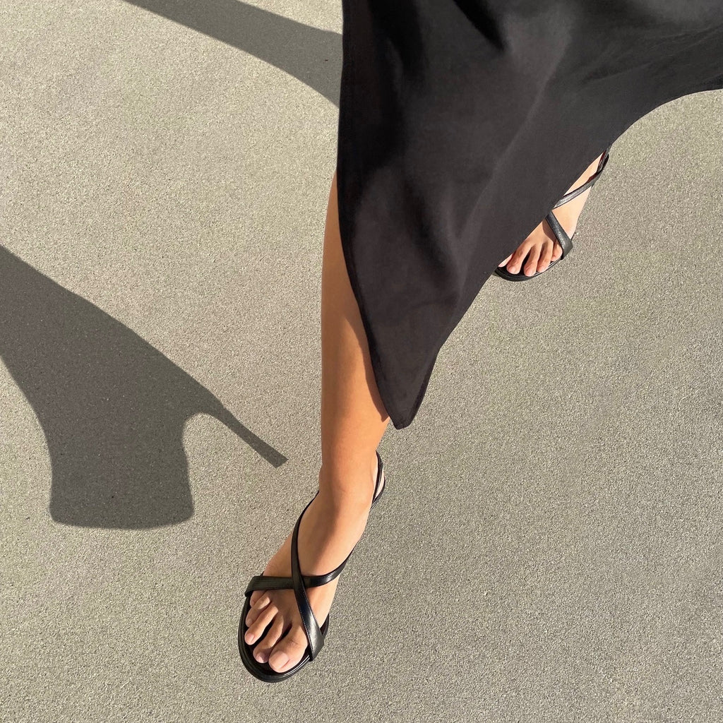 Neil J Rodgers Yin sandal in black nappa leather with a round toe, minimal criss cross instep straps, 60mm heel, and slingback strap is worn with a black long sleeve midi dress.