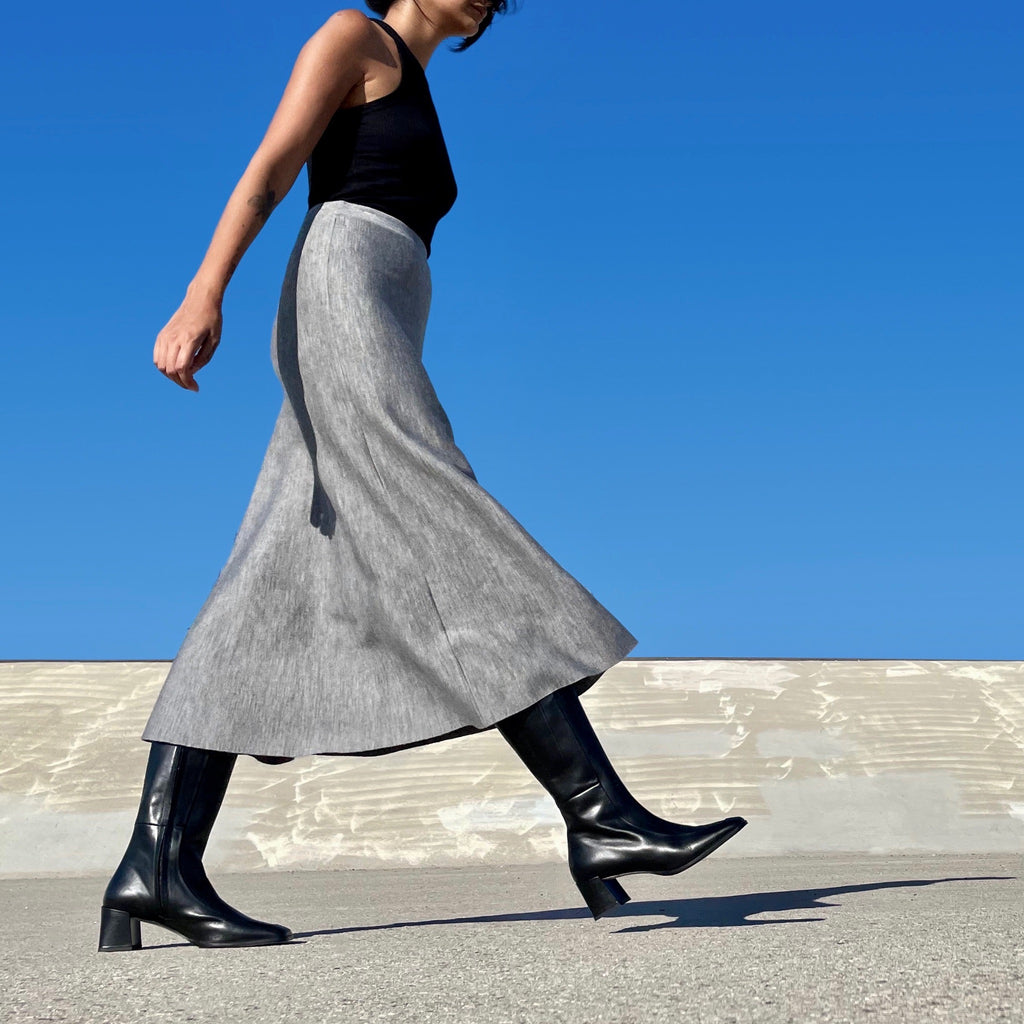 Neil J Rodgers Laura 90s knee boot with a rounded square toe, fitted leg that hits just below the knee, and comfortable block heel made from black Italian calf leather paired with a grey midi skirt.