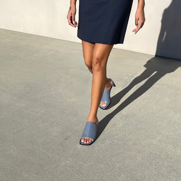 Neil J Rodgers Sue Mule in blue nappa leather with an exaggerated square toe, wide stretch nappa instep strap, padded footbed, and 60mm heel is worn with a navy mini dress.
